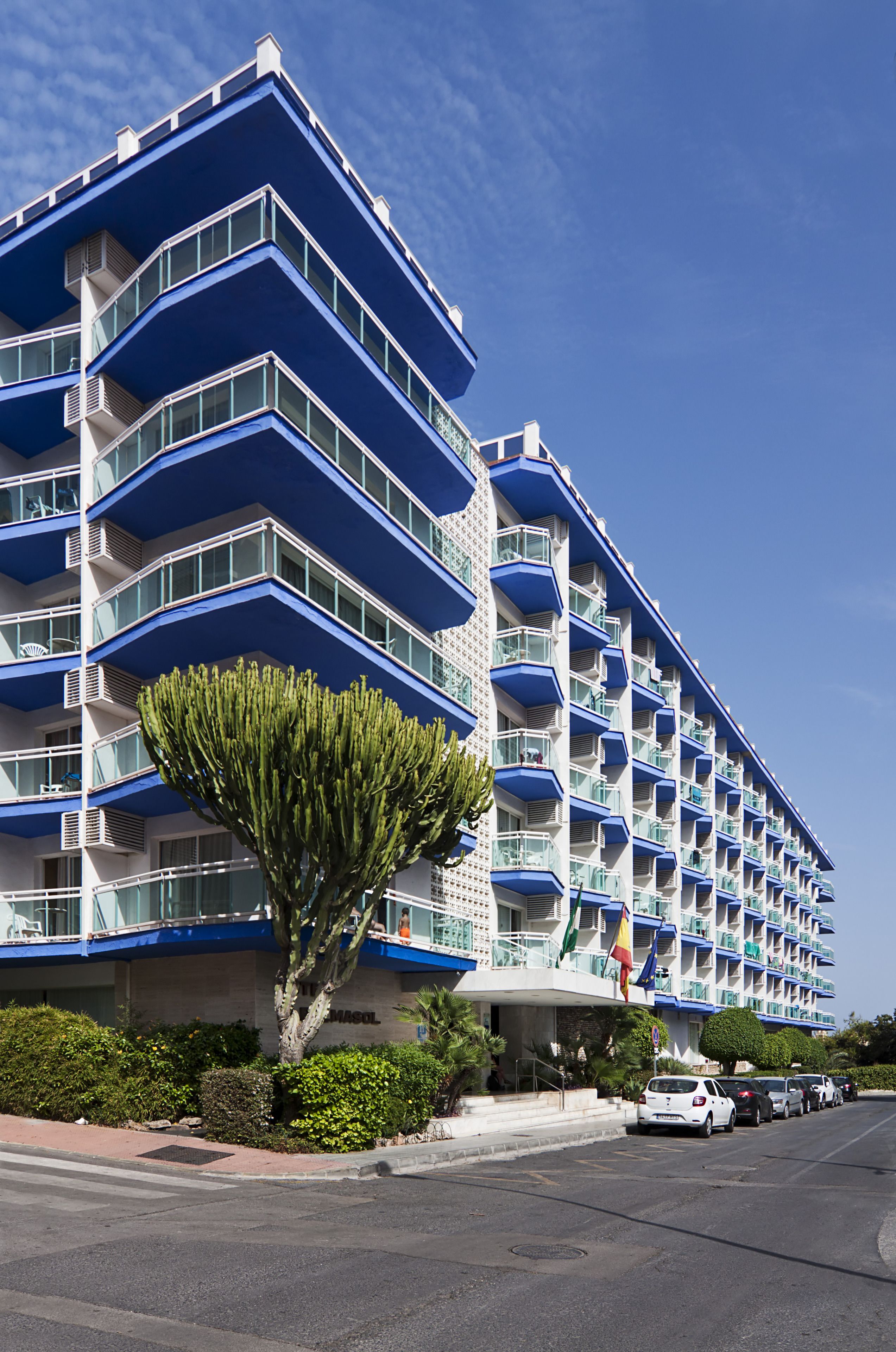 Un hotel de varios pisos con balcones, una bandera y un estacionamiento.