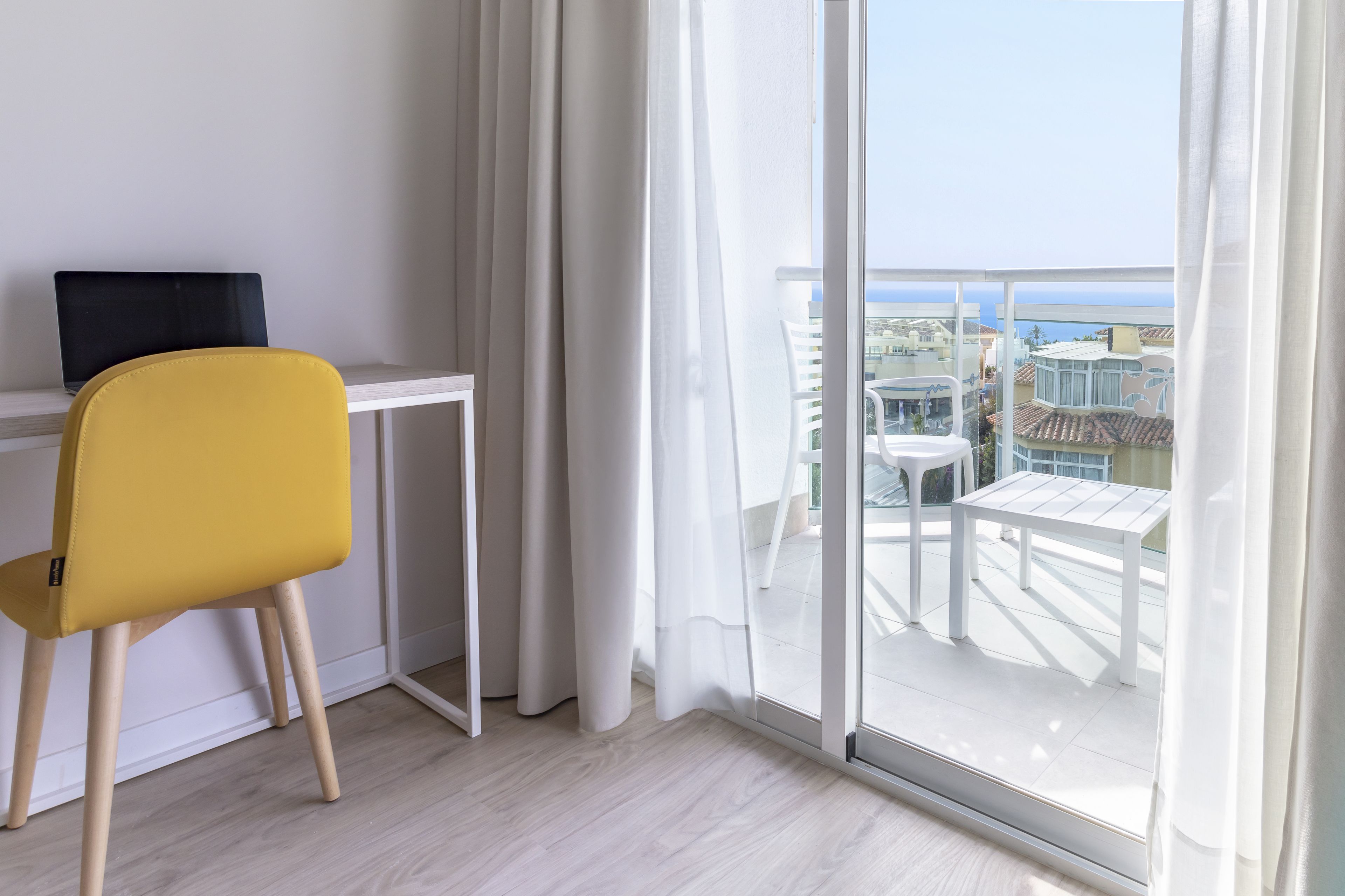 Ein modernes Zimmer mit einem gelben Sessel, einem weißen Schreibtisch und einer großen Schiebetür, die auf einen Balkon mit Gartenmöbeln führt.