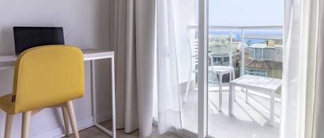 Ein modernes Zimmer mit einem gelben Sessel, einem weißen Schreibtisch und einer großen Schiebetür, die auf einen Balkon mit Gartenmöbeln führt.