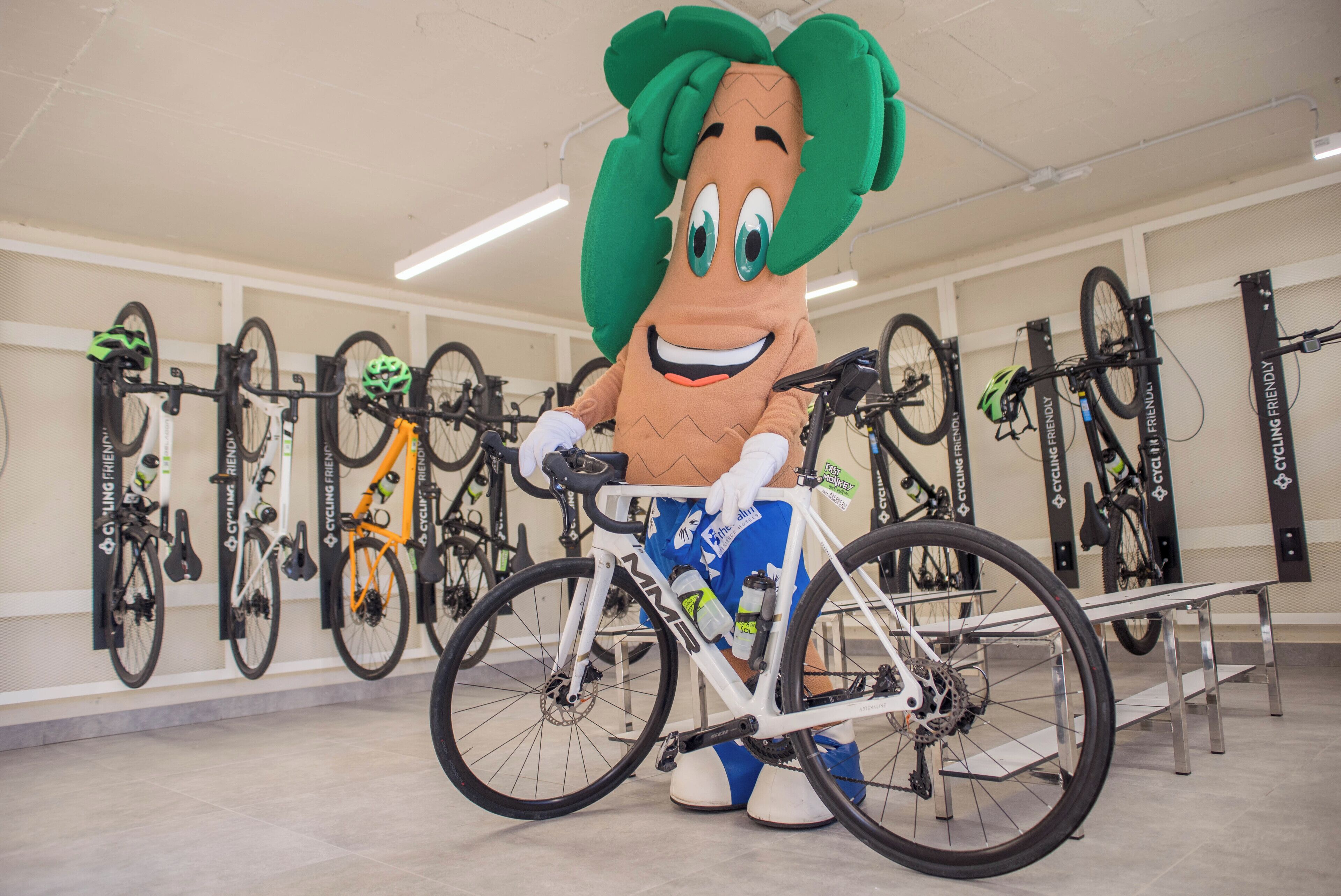 A mascot with a vegetable head standing next to a bicycle in a room with multiple bicycles mounted on the wall.