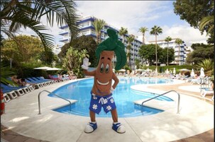 Eine Maskotte steht an einem Pool mit Palmen und einem Hotel im Hintergrund.