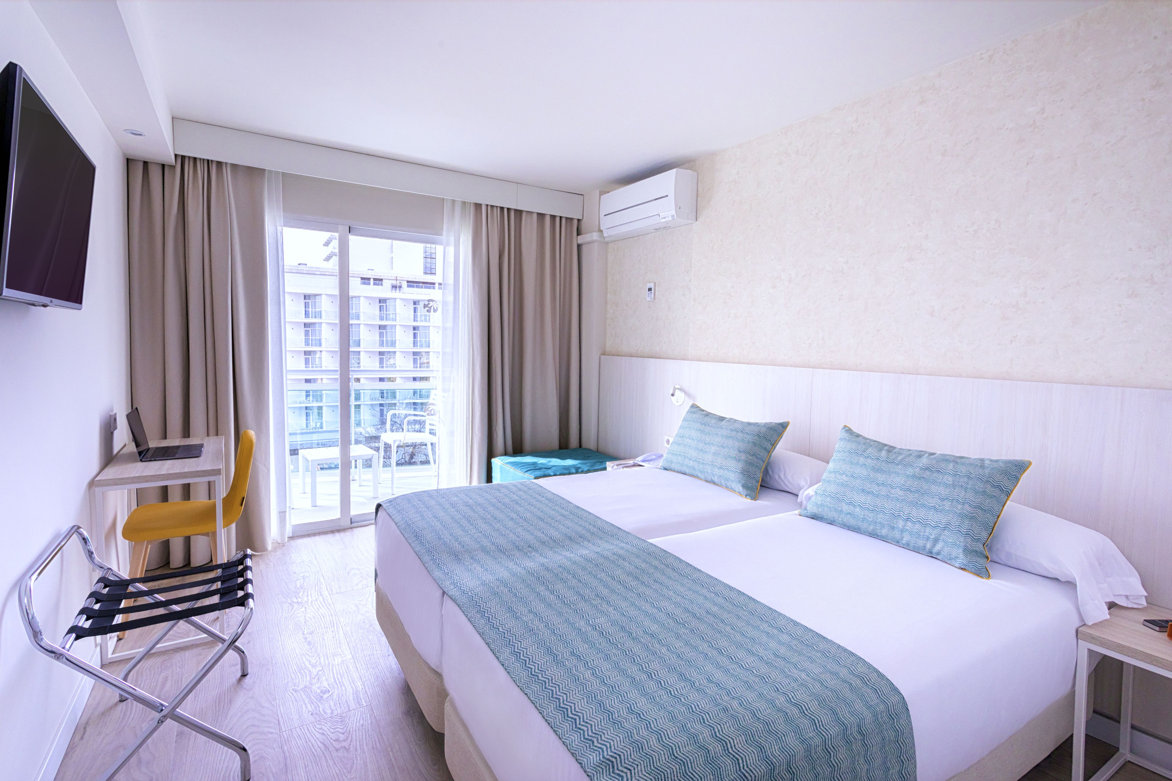 Ein Hotelzimmer mit Bett, Schreibtisch, Laptop, Stuhl, Nachttisch und Blick ins Freie.