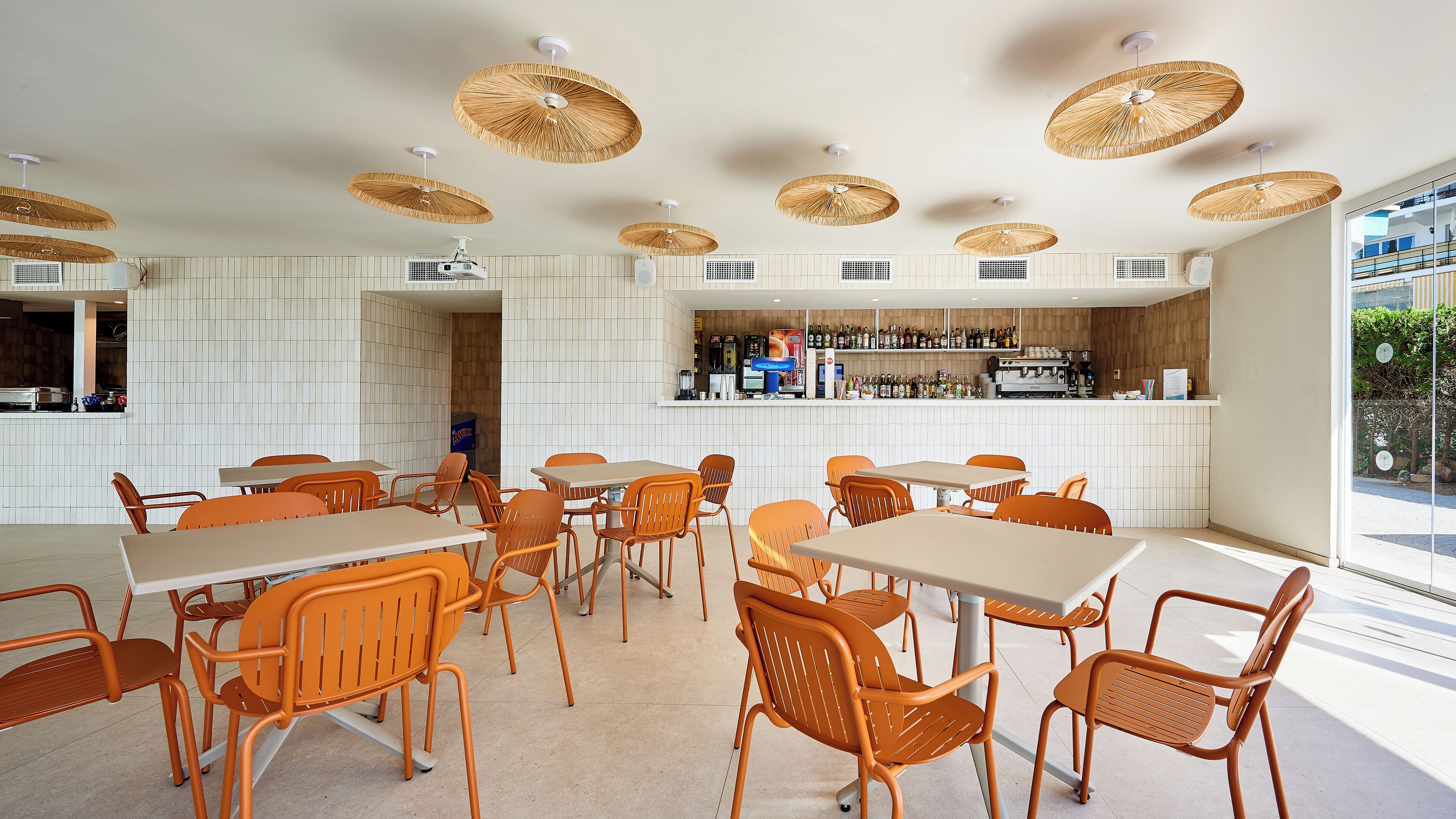 Een modern restaurantinterieur met witte betegelde muren, oranje stoelen en een bar met verschillende flessen en een koffieapparaat.