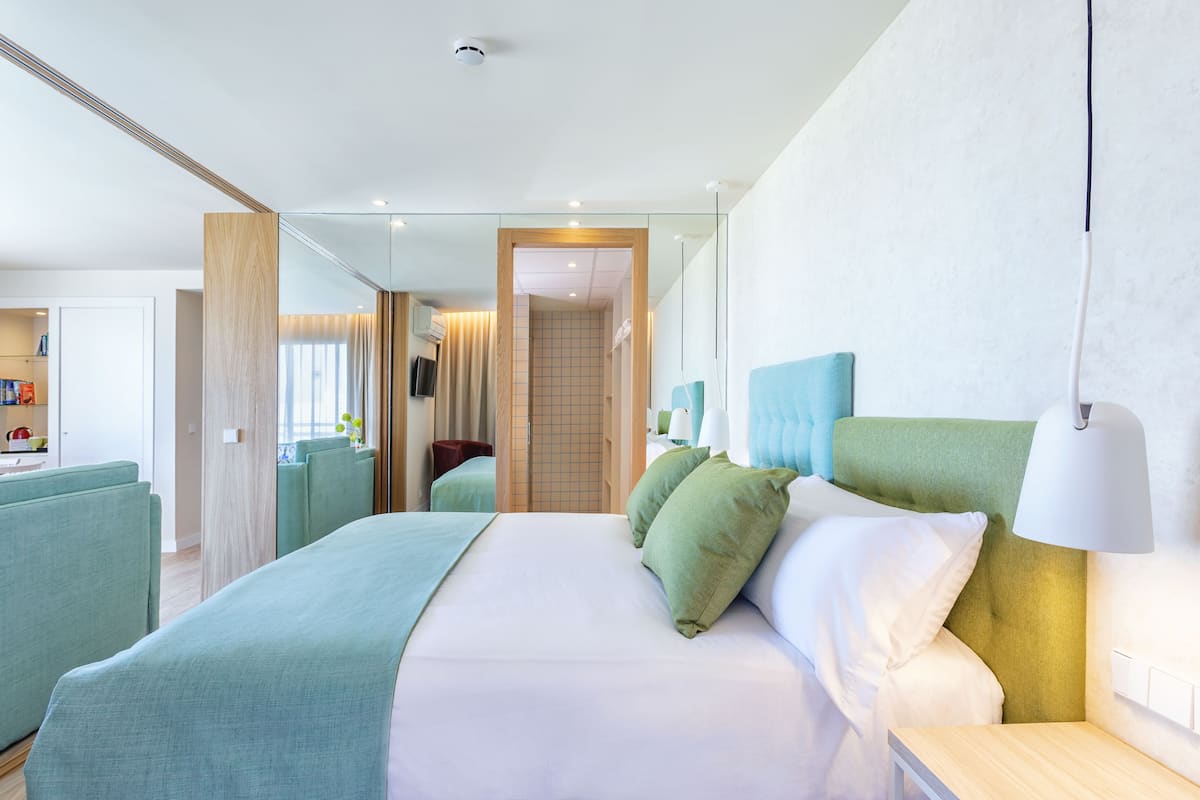 Una habitación de hotel moderna con una cama grande, ropa de cama verde y blanca, una mesita de noche con lámpara y vistas al pasillo.