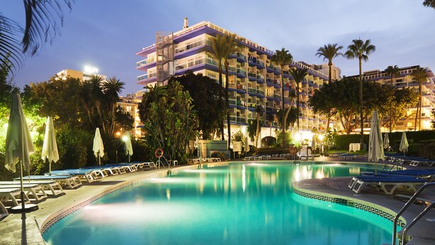 A hotel pool area with lounge chairs and umbrellas, surrounded by palm trees and a multi-story building with balconies. - Hotel Palmasol Puerto Marina (Benalmádena)
