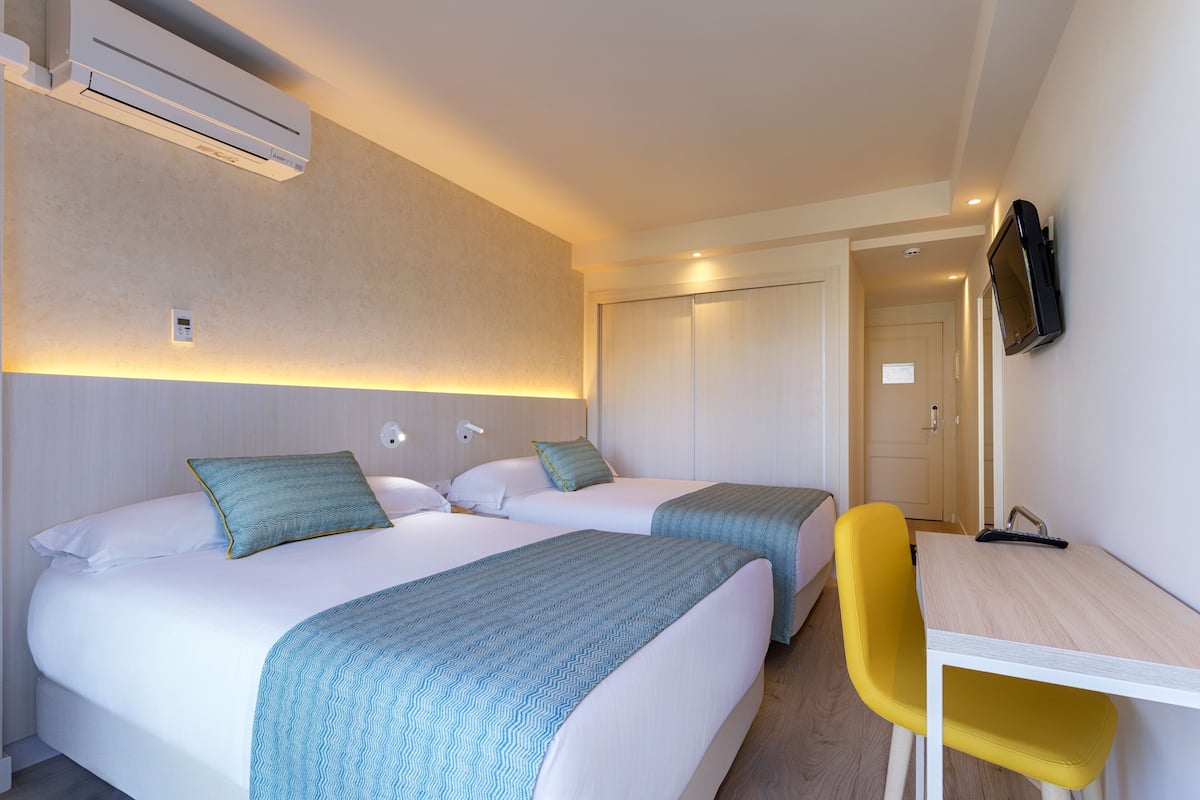 Habitación de hotel con dos camas, un escritorio, una silla, aire acondicionado y un televisor montado en la pared.