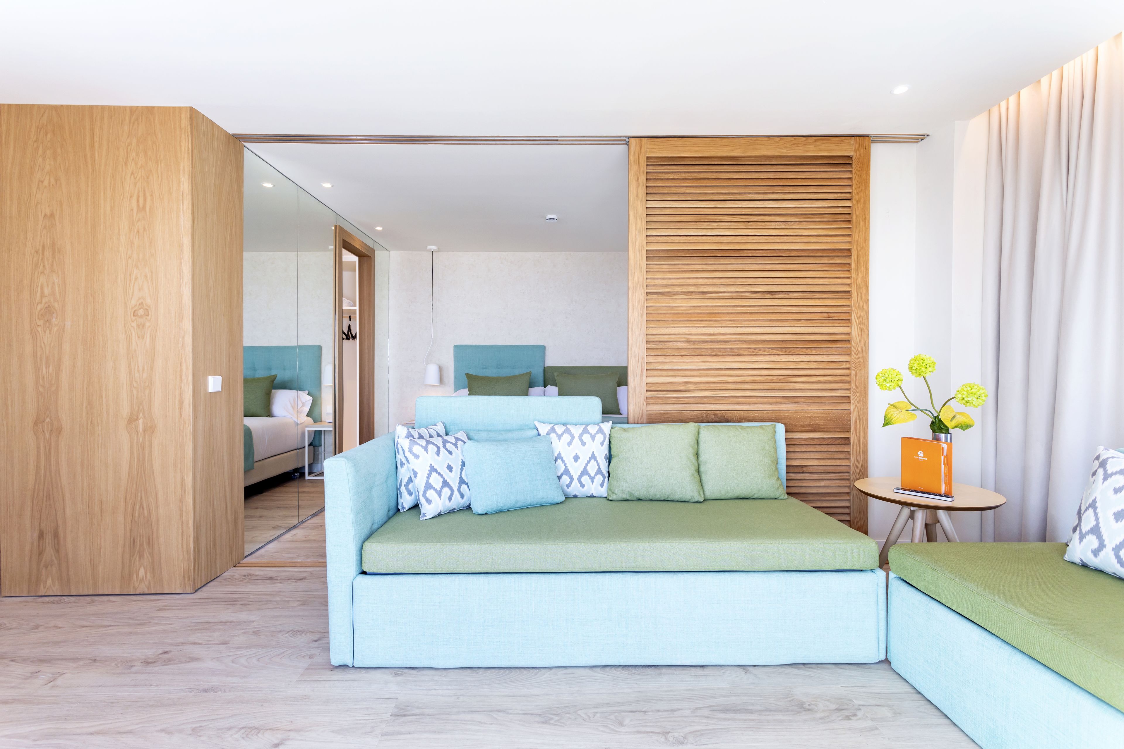 A modern hotel room with a green sofa, a wooden accent wall, and a small table with a vase of flowers.