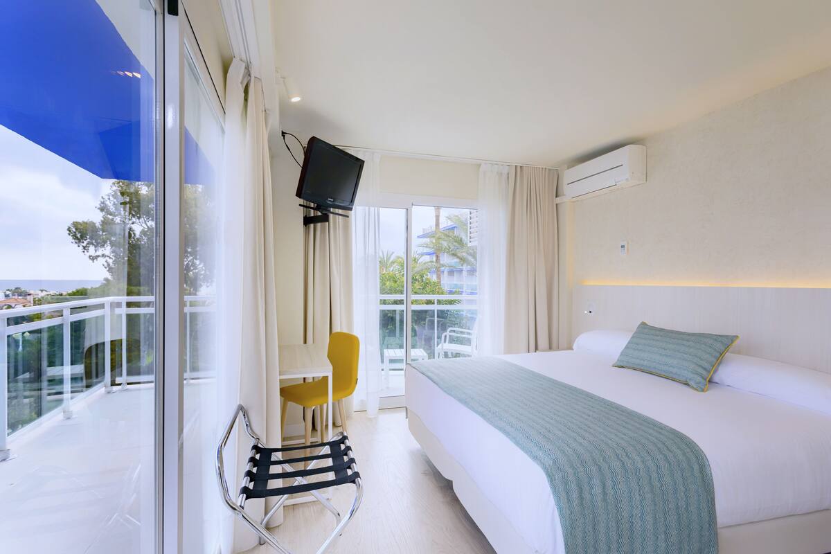 Habitación de hotel con cama, televisión, balcón con una silla y vistas al exterior.
