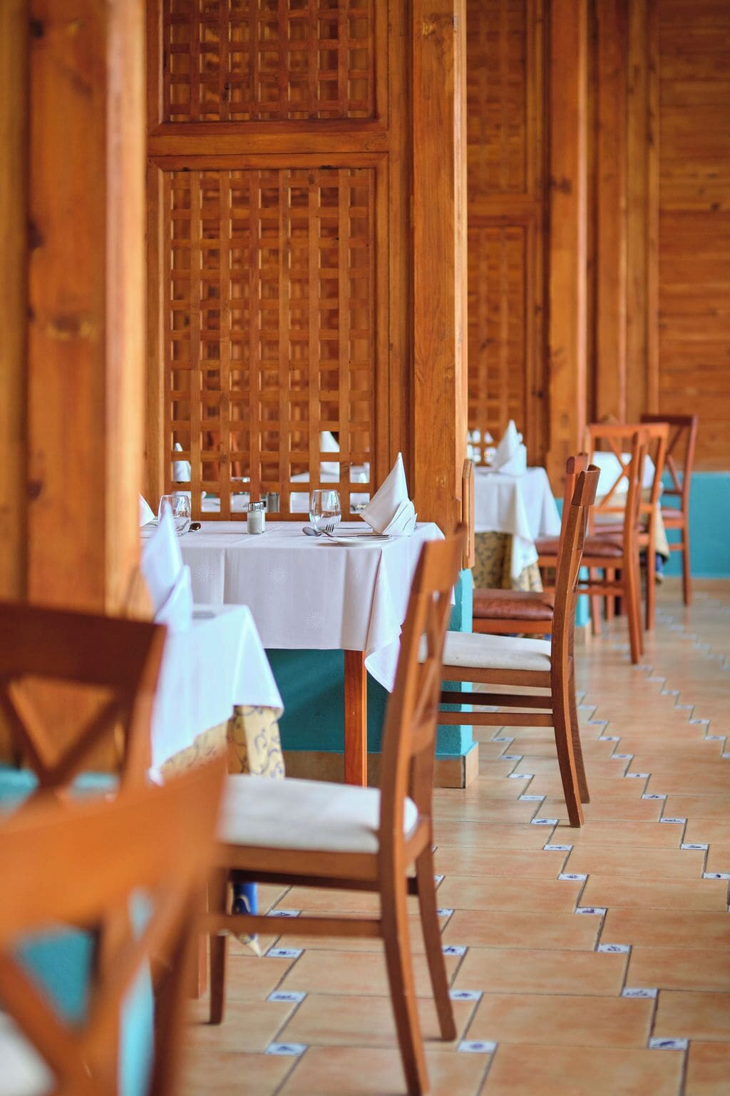 A row of wooden tables with white tablecloths, wooden chairs, and a patterned floor.