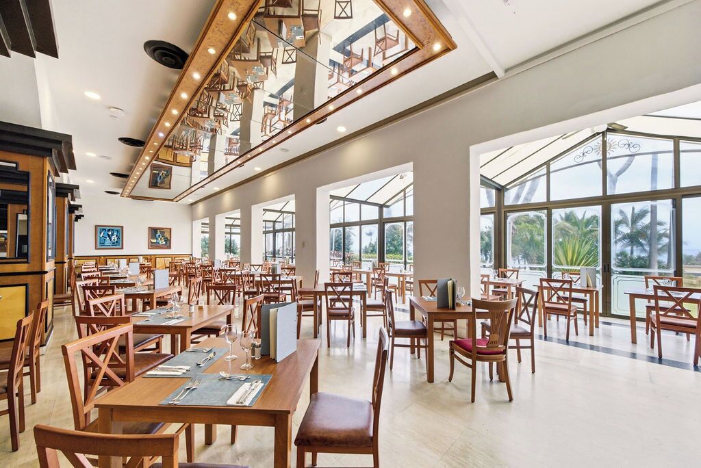 Uma ampla sala de jantar com várias mesas e cadeiras, grandes janelas que oferecem vista para palmeiras e um design de teto moderno com iluminação integrada.