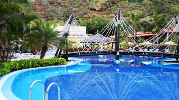 A swimming pool with a rope bridge, palm trees, and a mountainous backdrop.