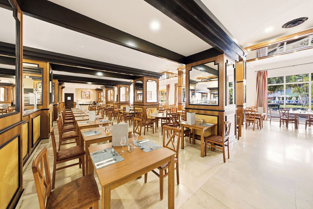 Un restaurante amplio con numerosas mesas y sillas, vigas de madera en el techo y grandes ventanales que dejan entrar luz natural.