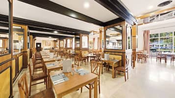 A spacious restaurant with multiple dining tables and chairs, wooden beams on the ceiling, and large windows allowing natural light.