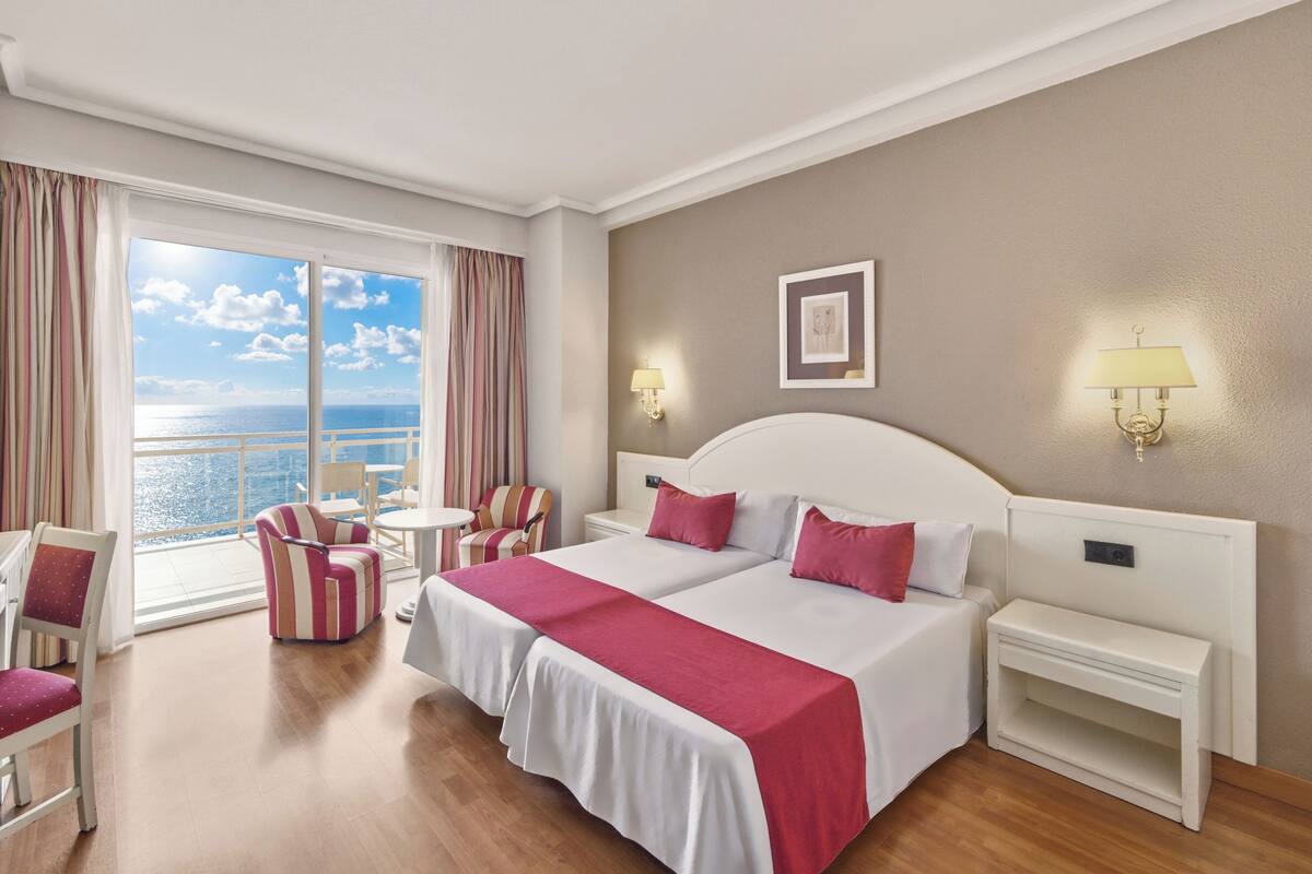 A hotel room with two beds, a view of the sea, a chair, a small table, and two nightstands with lamps.