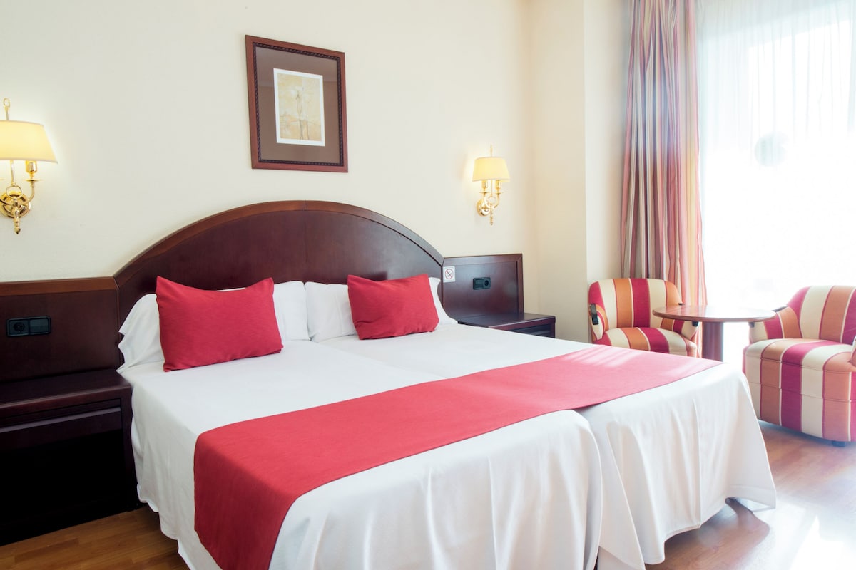 Habitación de hotel con una cama grande, dos mesitas de noche, un tocador, una silla, una pequeña mesa y un ventanal con cortinas.