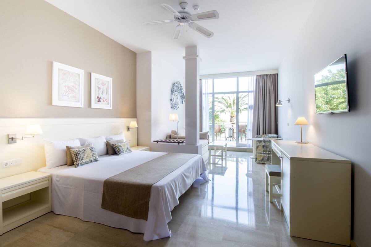 A hotel room with a large bed, a desk, a chair, a TV, and a balcony with a view.
