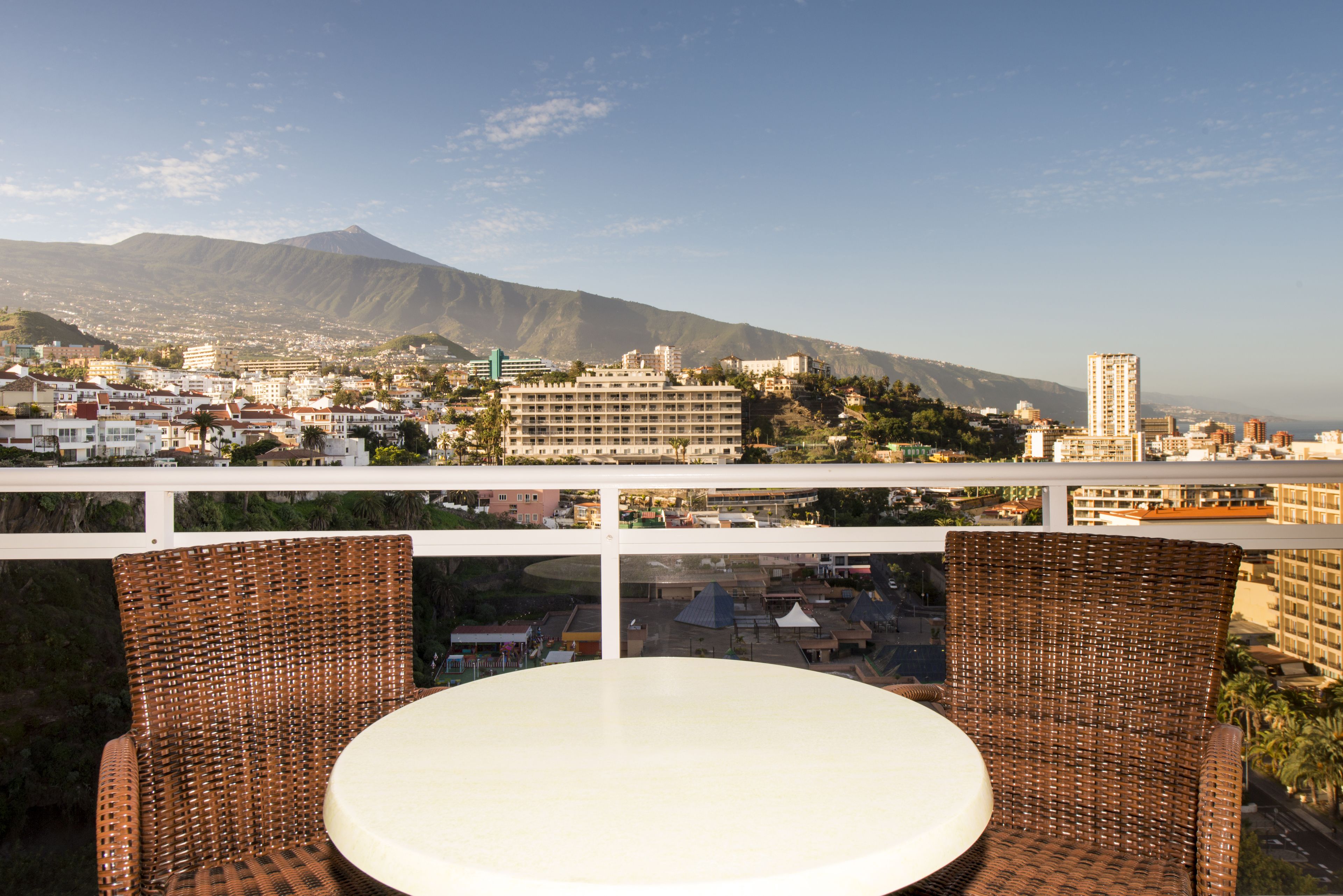 A balcony with wicker chairs and a table overlooking a cityscape with buildings and a mountain.