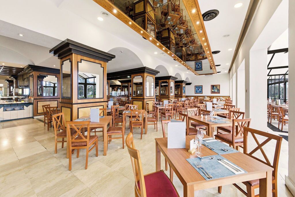 Un restaurante espacioso con mesas y sillas de madera, techos altos con vigas a la vista y ventanas grandes que dejan entrar luz natural.