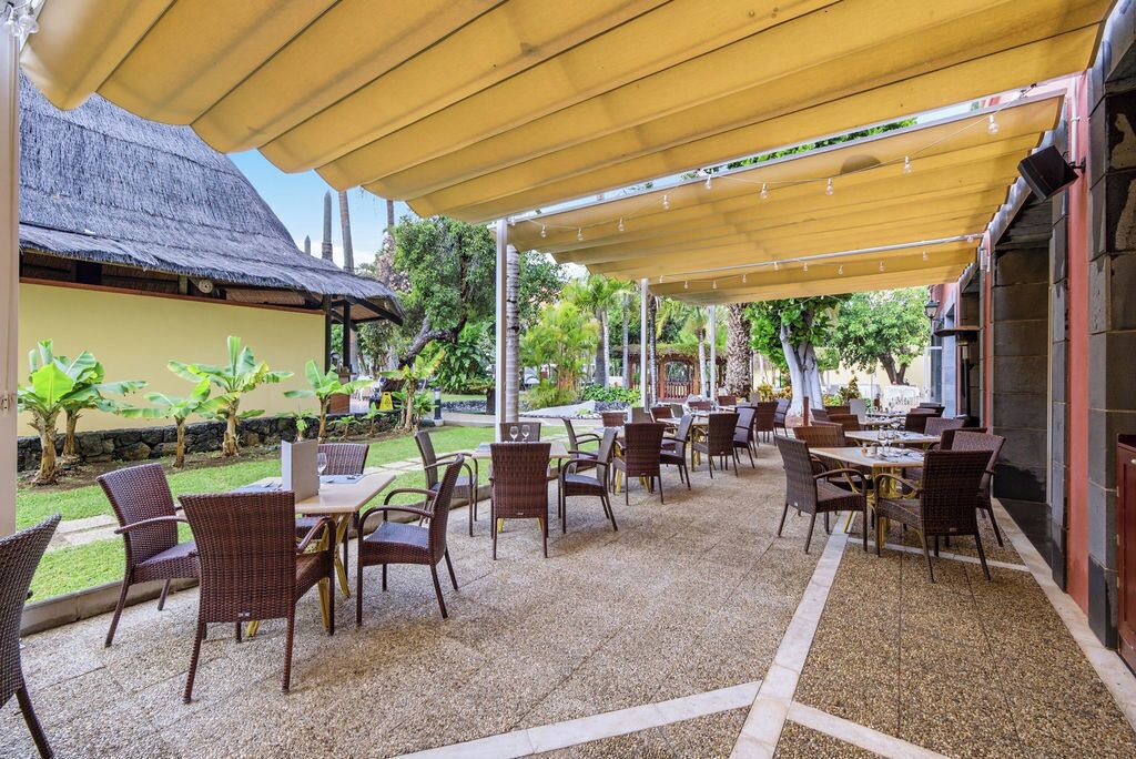 An outdoor patio area with tables and chairs, a covered seating area, and a building with a thatched roof in the background.