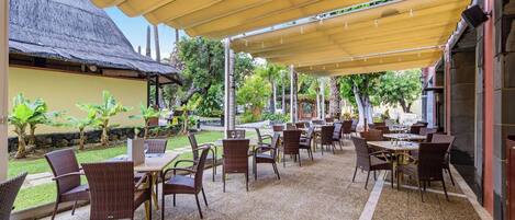An outdoor patio area with tables and chairs, a covered seating area, and a building with a thatched roof in the background.