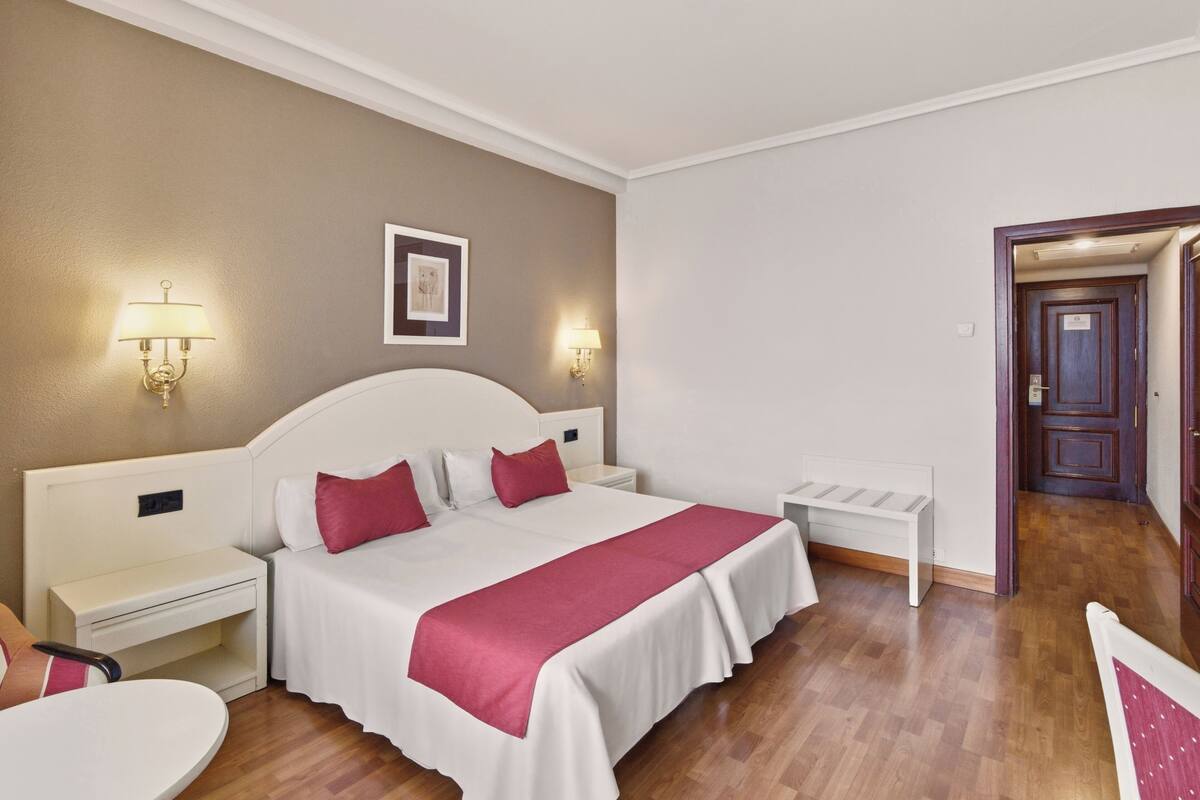 A hotel room with a large bed, bedside tables, a chair, and a door leading to another room.