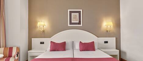 A double bed with white and red bedding, two bedside tables with lamps, a framed picture on the wall, and a striped armchair.
