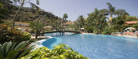 A large outdoor swimming pool surrounded by lush greenery and palm trees, with a bridge crossing over the pool.