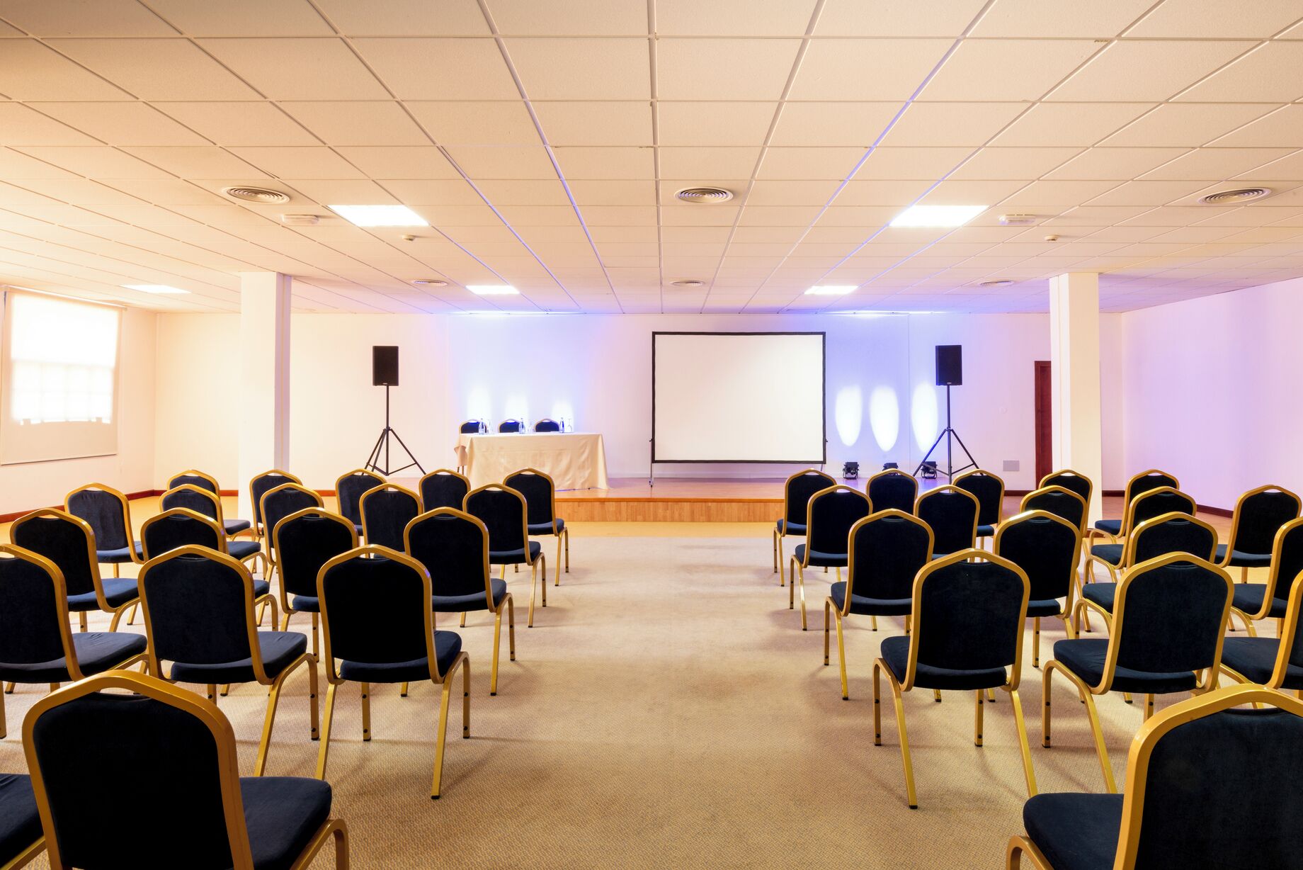 Un salón de conferencias con filas de sillas, un atril, una pantalla de proyector y altavoces.