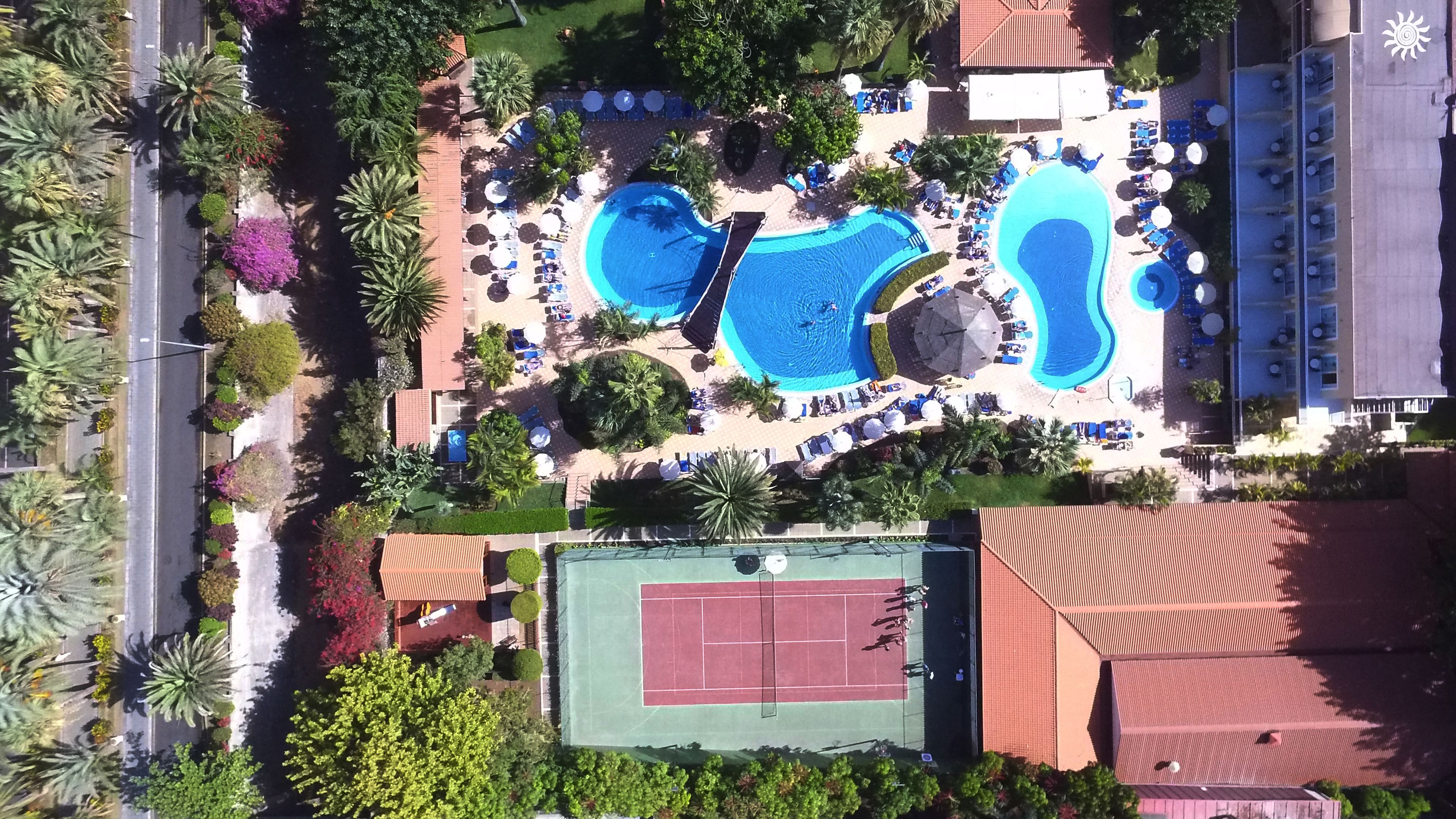 Vista aèria d'un complex turístic amb diverses piscines, gandules i una pista de tennis.