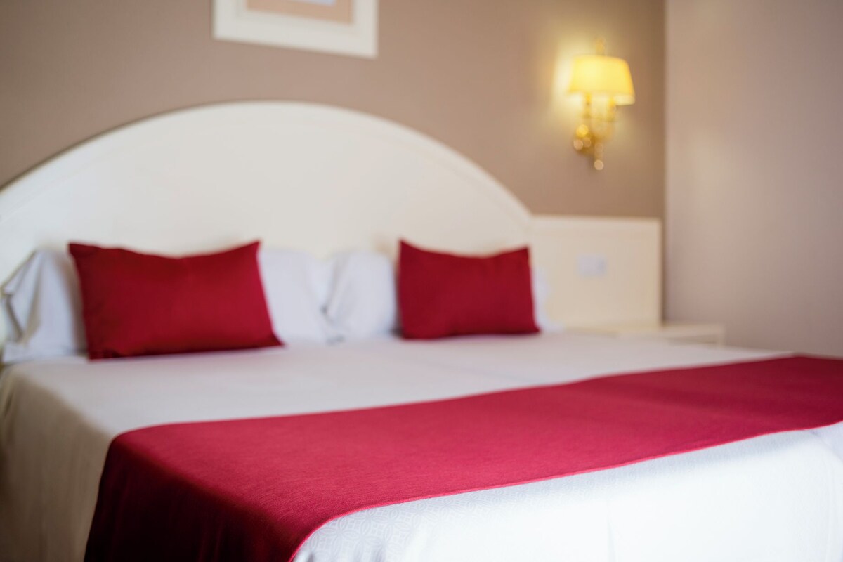 Una cama con cabecera blanca y almohadas rojas, una colcha blanca y una lámpara de pared.