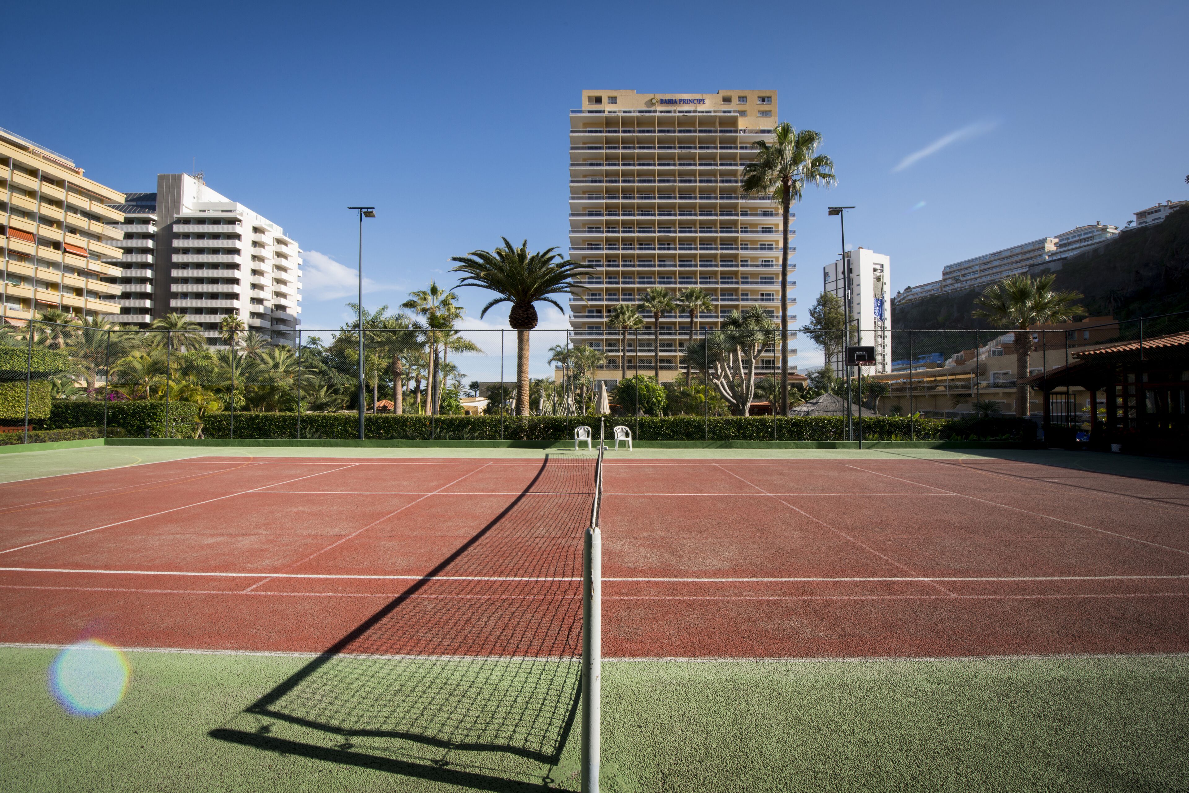 A tennis court with a net, surrounded by palm trees and a hotel building in the background.