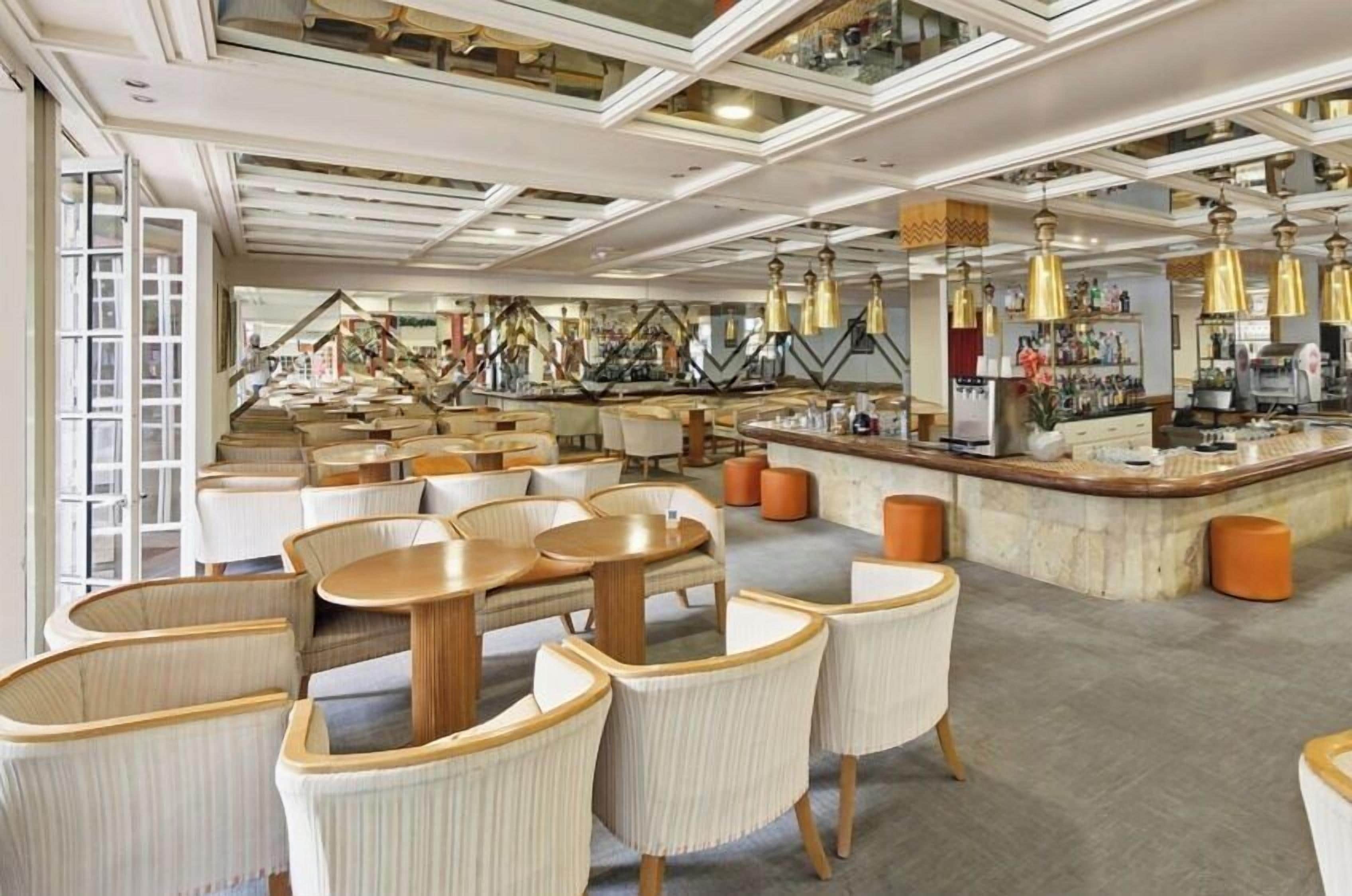 A modern restaurant with round tables and cushioned chairs, a bar area, and a view of the city through large windows.