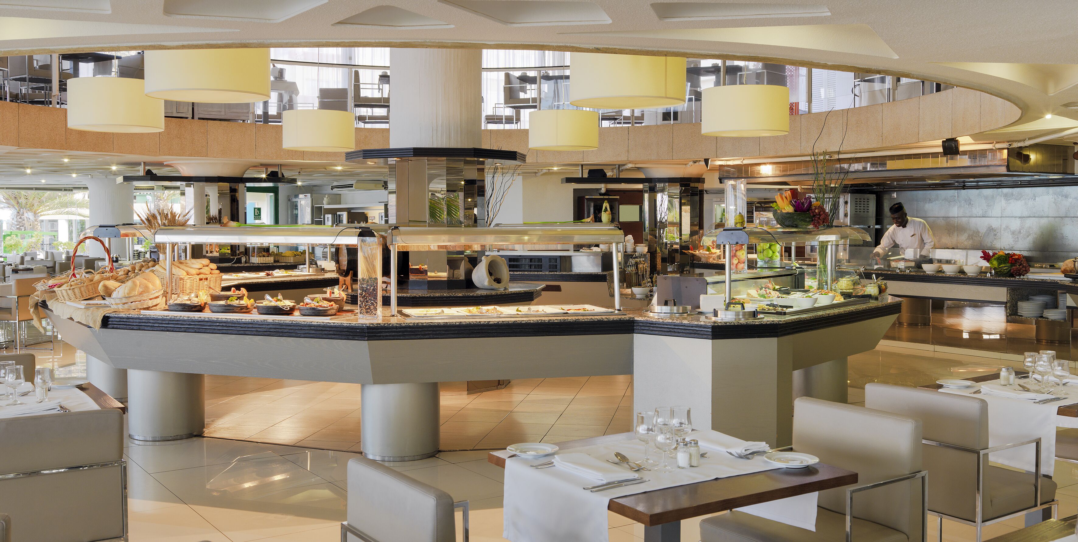 A modern hotel lobby with a central food display, multiple seating areas, and a high ceiling with large windows.
