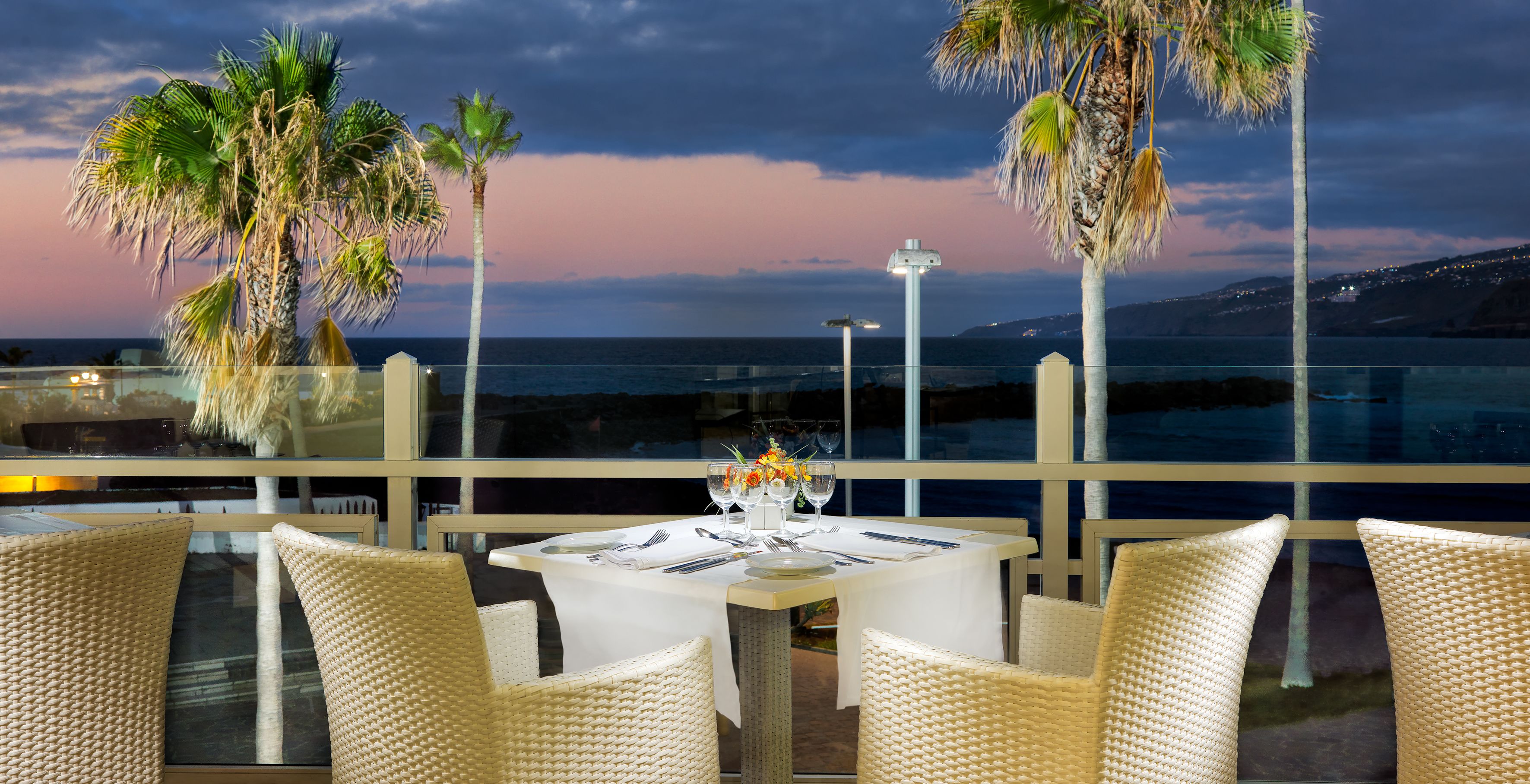 A dining table set for two with wicker chairs, overlooking a coastal view with palm trees and a sunset.