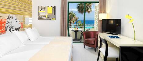 A hotel room with a bed, a desk, a TV, and a view of the beach.