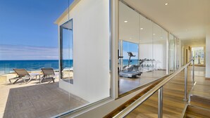 Un interior moderno con un muro de vidrio con vista al mar, una terraza de madera con sillones de descanso y una caminadora al fondo.