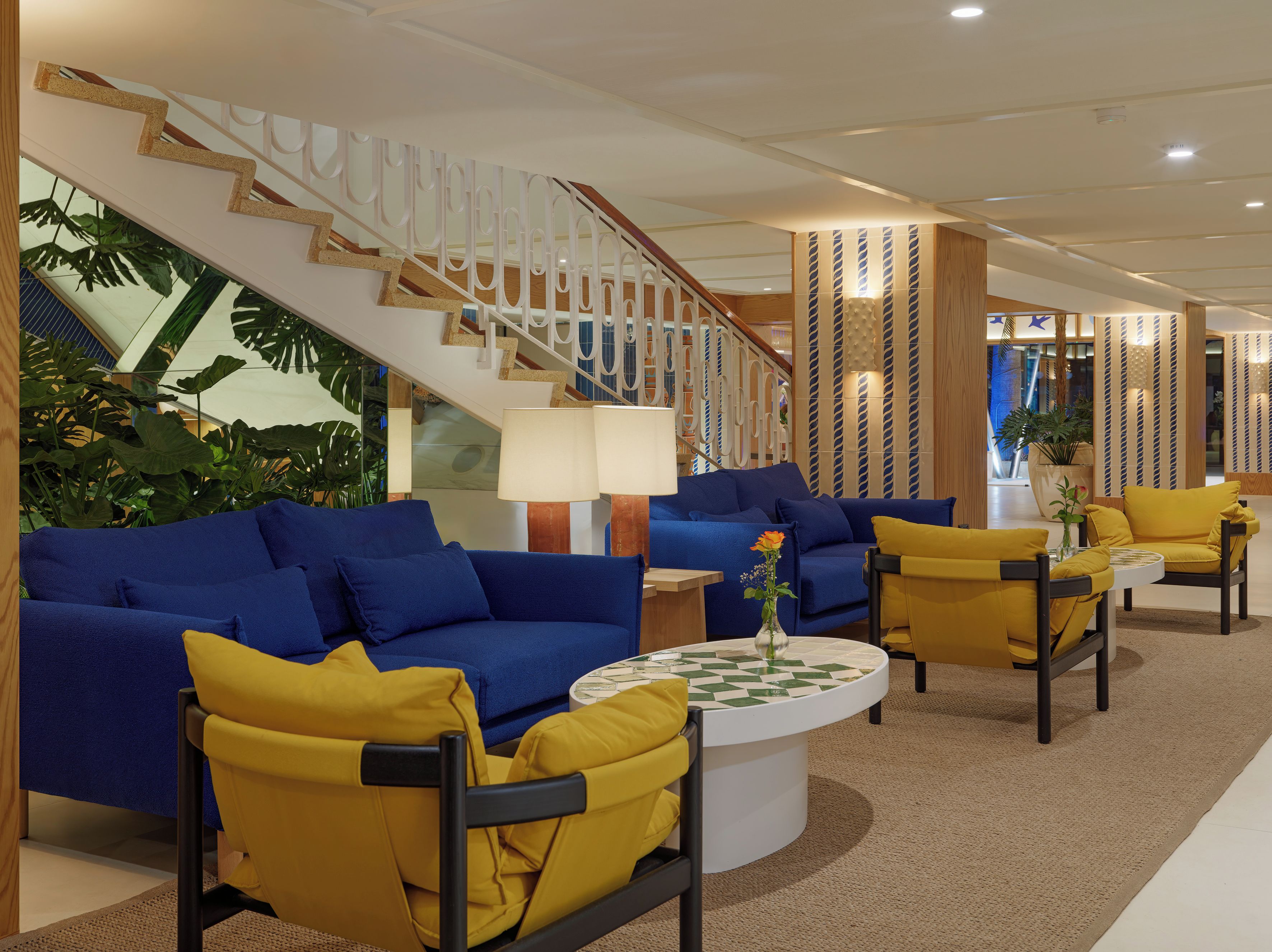 Un hall d’entrée moderne avec des canapés bleus, des fauteuils jaunes, un escalier et une grande plante.