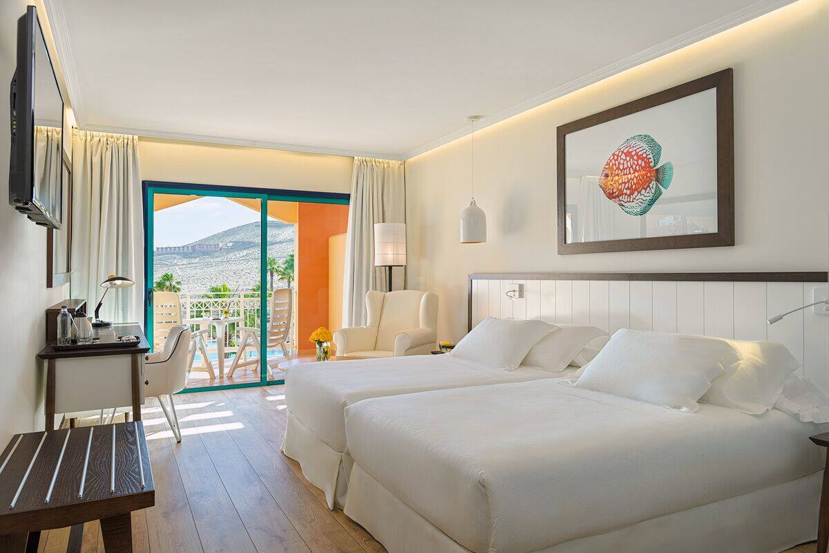 Une chambre d’hôtel avec deux lits, un bureau, une chaise et un balcon donnant sur une montagne.