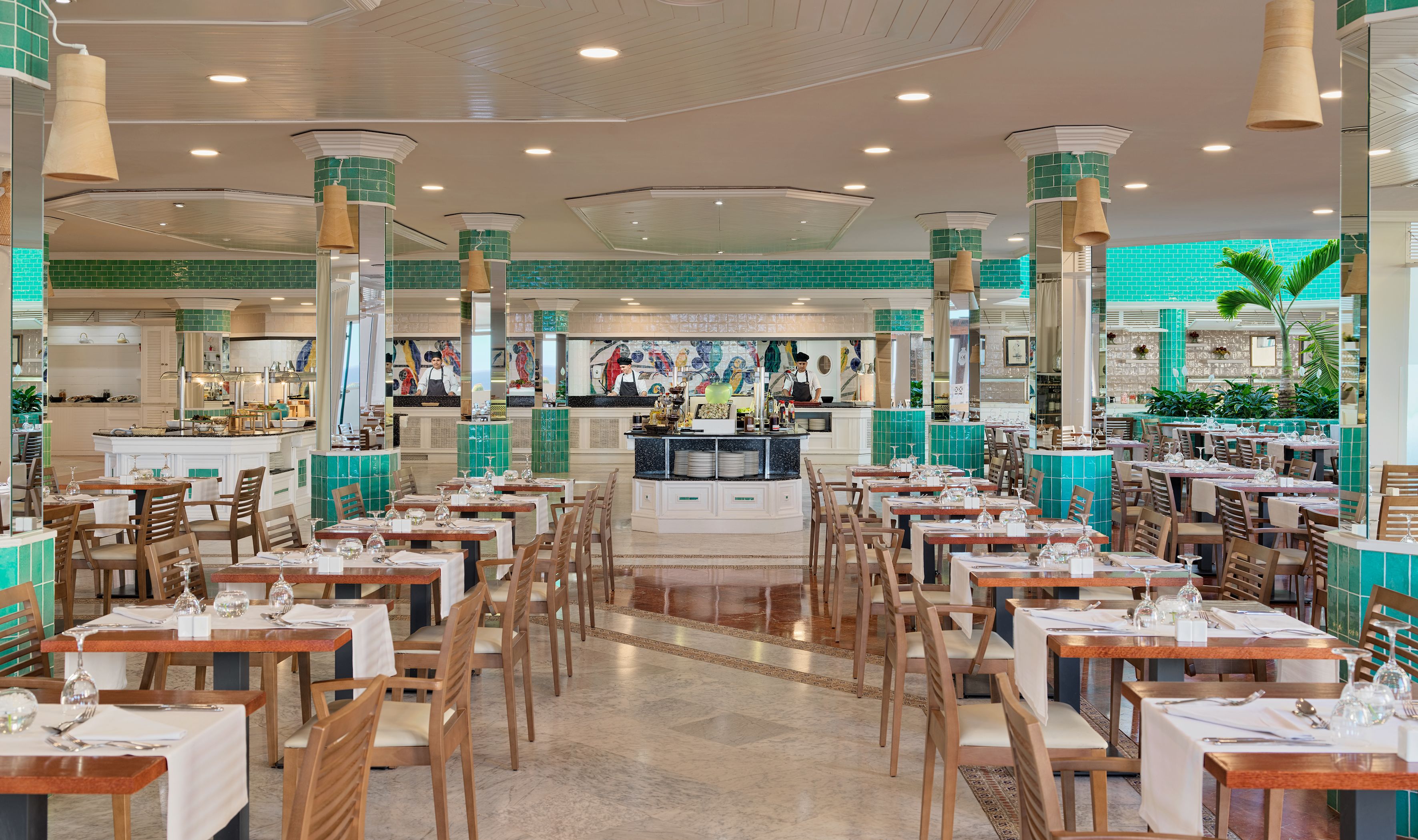 A spacious restaurant with multiple dining tables and chairs, a central bar area, and a tiled green wall.