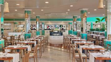 A spacious restaurant with multiple dining tables and chairs, a central bar area, and a tiled green wall.
