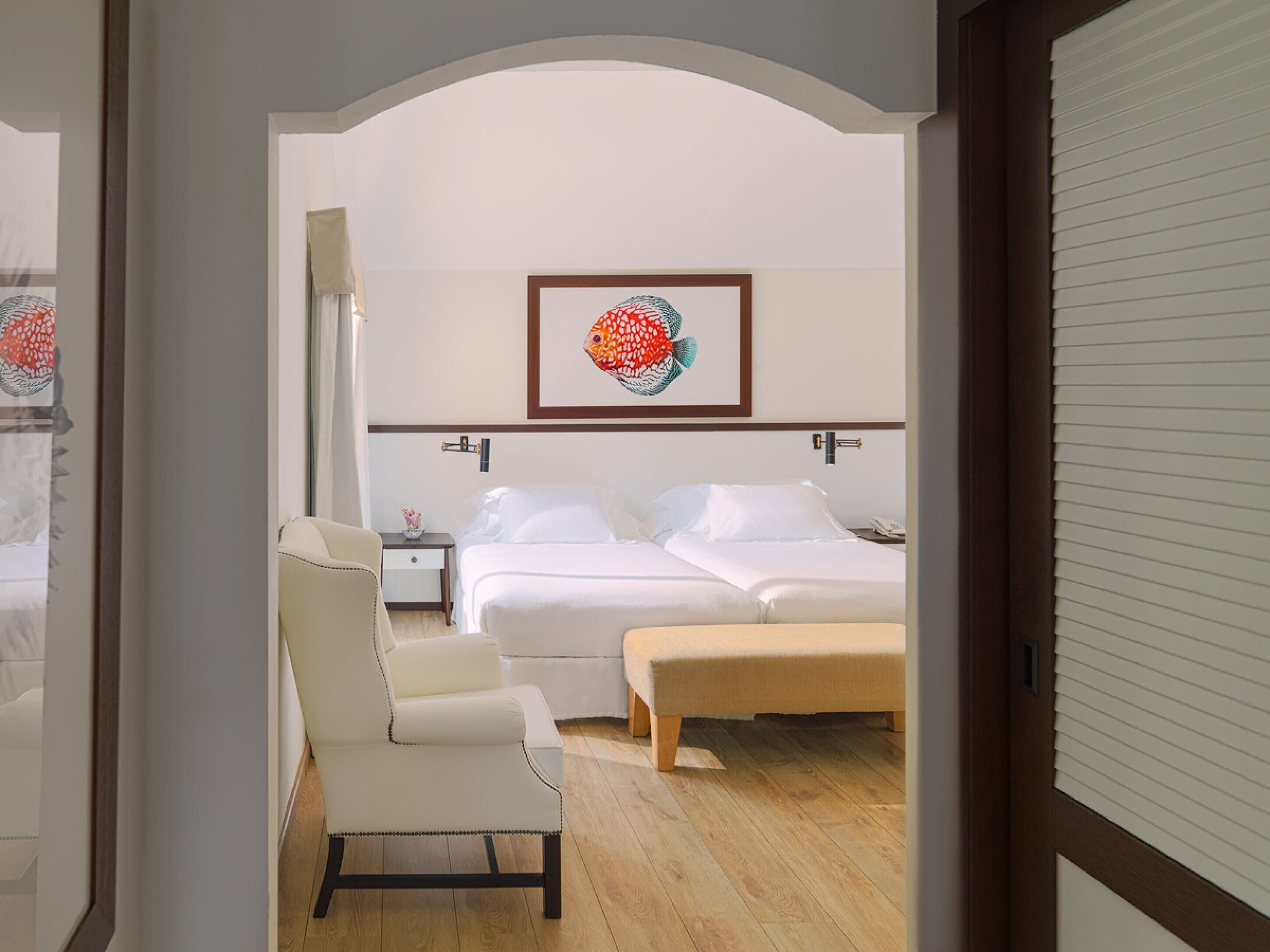 A hotel room with a bed, a chair, a bedside table, and a framed artwork on the wall.