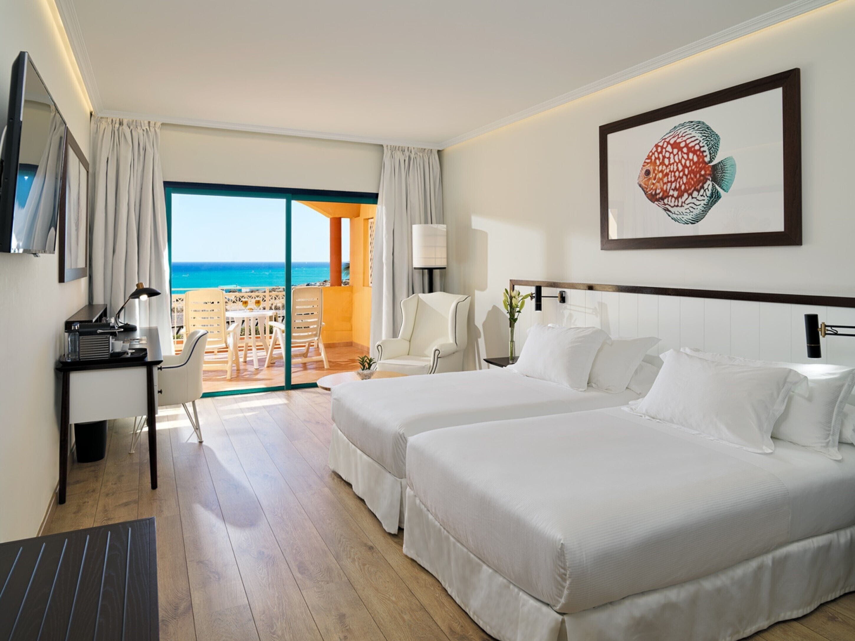A hotel room with two beds, a desk, a chair, a TV, and a view of the ocean.