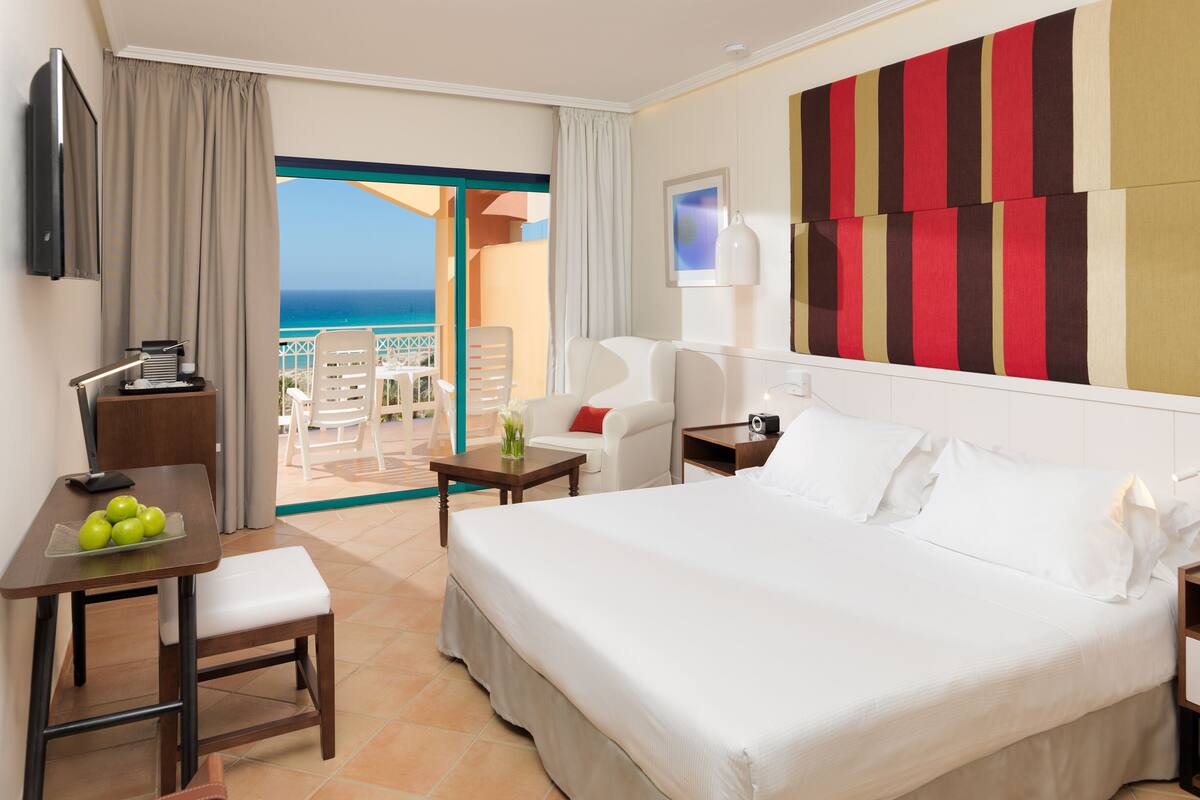 Une chambre d’hôtel avec un lit, un bureau avec une lampe, une chaise, une petite table avec un vase et une vue sur l’océan.