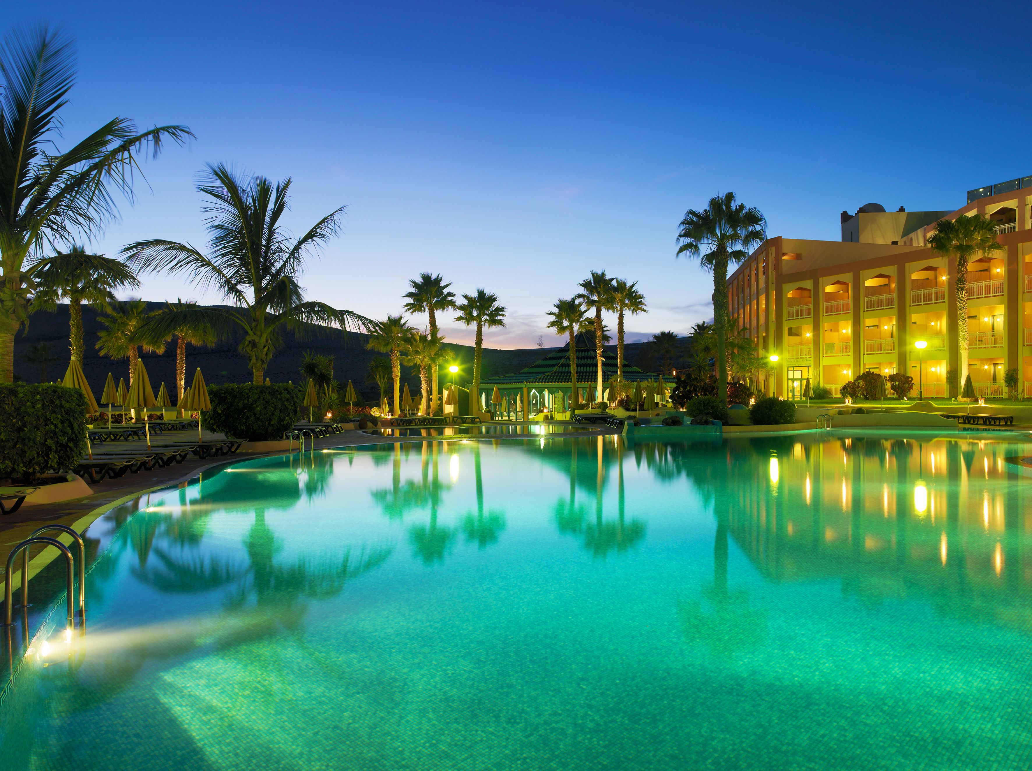A large outdoor swimming pool with clear water, surrounded by palm trees and a hotel building with multiple balconies.