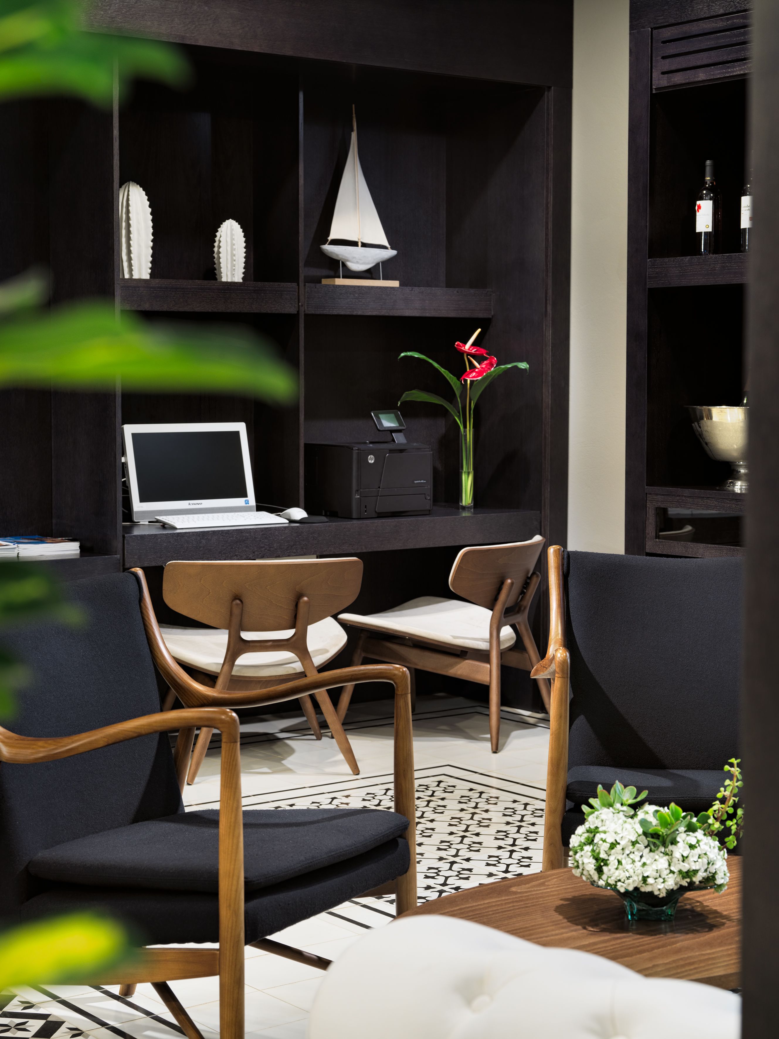 A modern living room with a dark wood bookshelf, a laptop, two wooden chairs, a white sofa, and a small table with a vase of flowers.