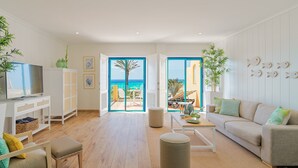 Een ruime woonkamer met een houten vloer, een bank, een televisie en uitzicht op zee door open deuren.