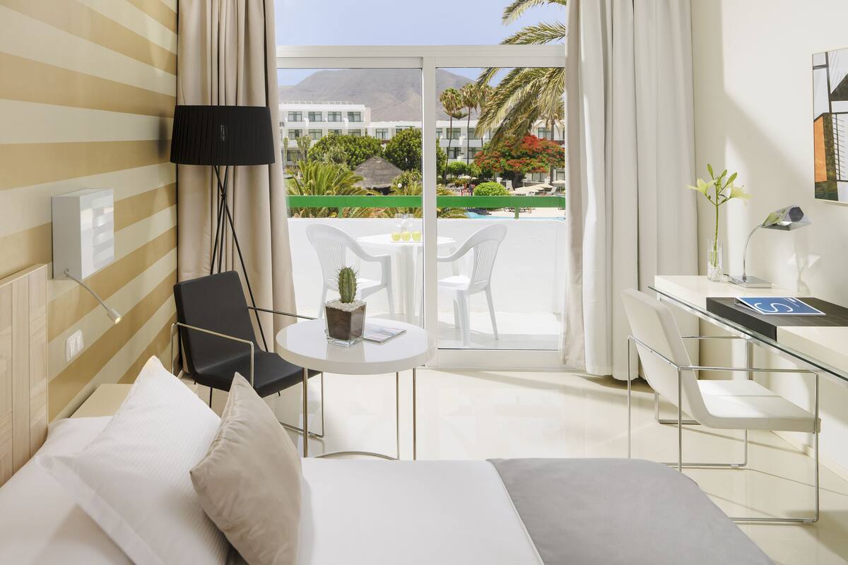 A hotel room with a bed, a desk, a chair, a small table with a cactus, and a view of a garden and buildings outside.