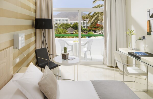 A hotel room with a bed, a desk, a chair, a small table with a cactus, and a view of a garden and buildings outside. - H10 Lanzarote Princess (Yaiza)