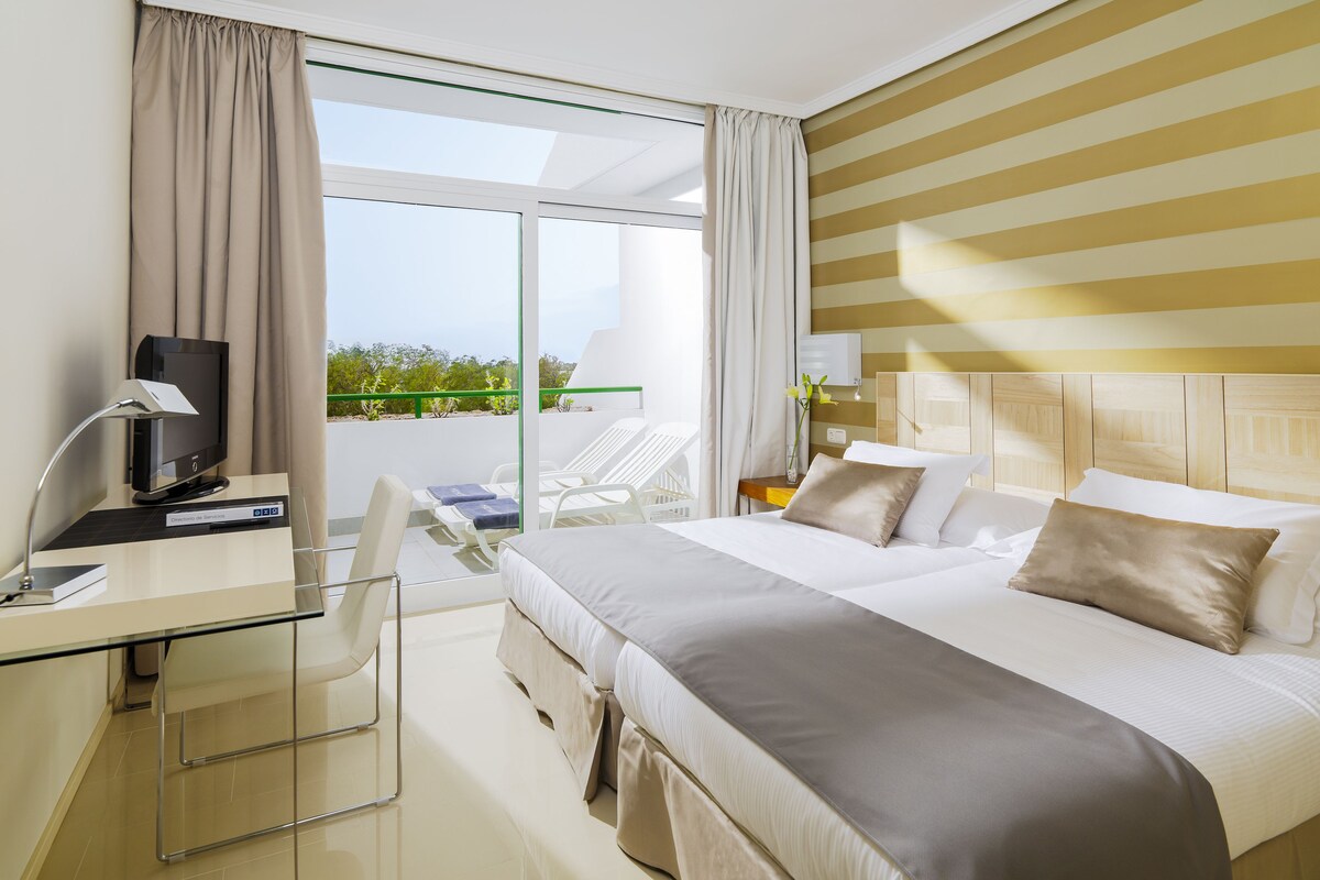 A hotel room with a bed, desk, chair, and a view of greenery outside.