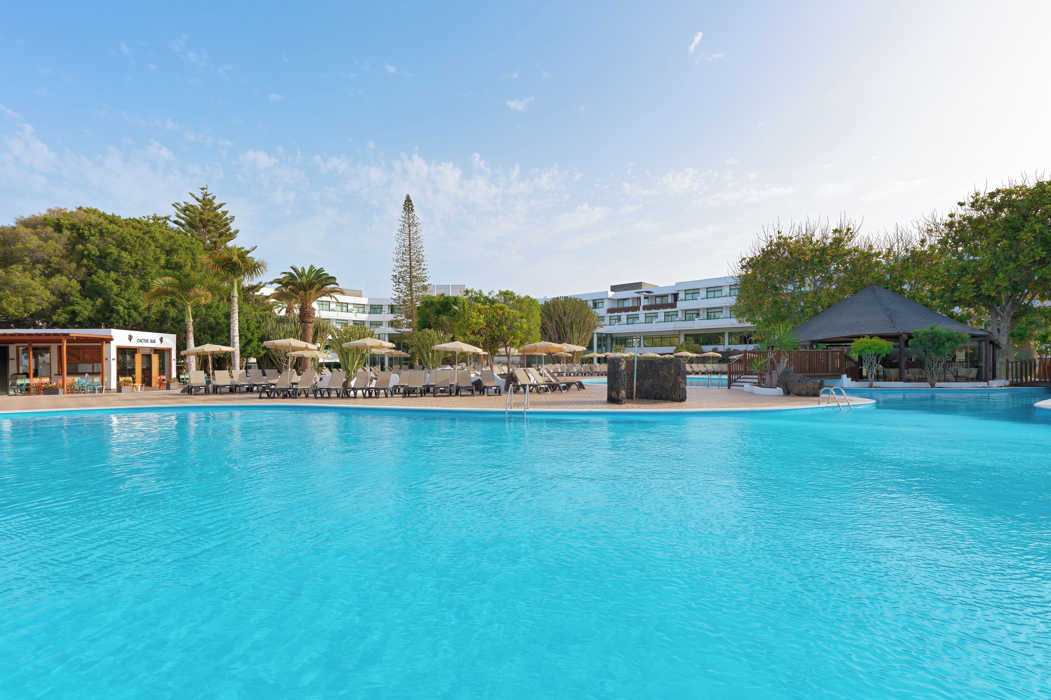 A clear blue pool with lounge chairs and umbrellas, surrounded by trees and buildings.